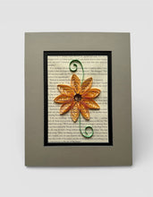 Load image into Gallery viewer, A framed artwork featuring a vibrant orange paper quilt flower resting on a page of text from a book.
