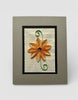 A framed artwork featuring a vibrant orange paper quilt flower resting on a page of text from a book.